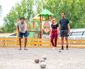 Familie spielt Boule auf dem Campingplatz, im Hintergrund Spielplatz mit Rutsche und Klettergerüst, Bäume und Wohnwagenzaun.