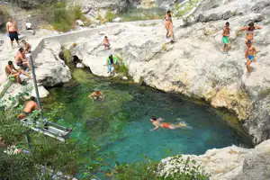 Naturpool zwischen Felsen mit mehreren Badegästen, Sprungbereich und Leiter am Rand, umgeben von Steinen und Pflanzen.