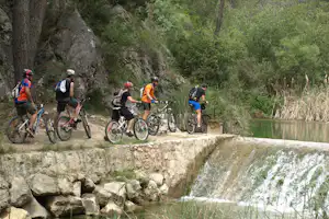 Radfahrer mit Helmen auf einem Schotterweg am Fluss, neben einem kleinen Wasserfall und grünem Uferbewuchs.