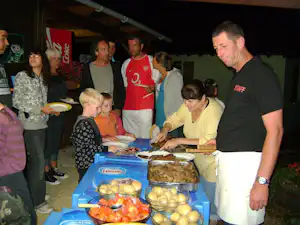 Grillbuffet mit mehreren Schüsseln und Tellern, Campingpersonal serviert am Tisch in einer überdachten Gemeinschaftsfläche.