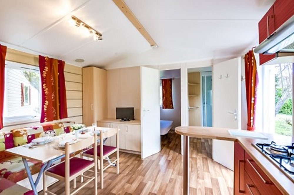 Innenansicht eines Wohnwagens mit Küchenzeile, Esstisch und Sitzbank, großen Fenstern und Holzfußboden.