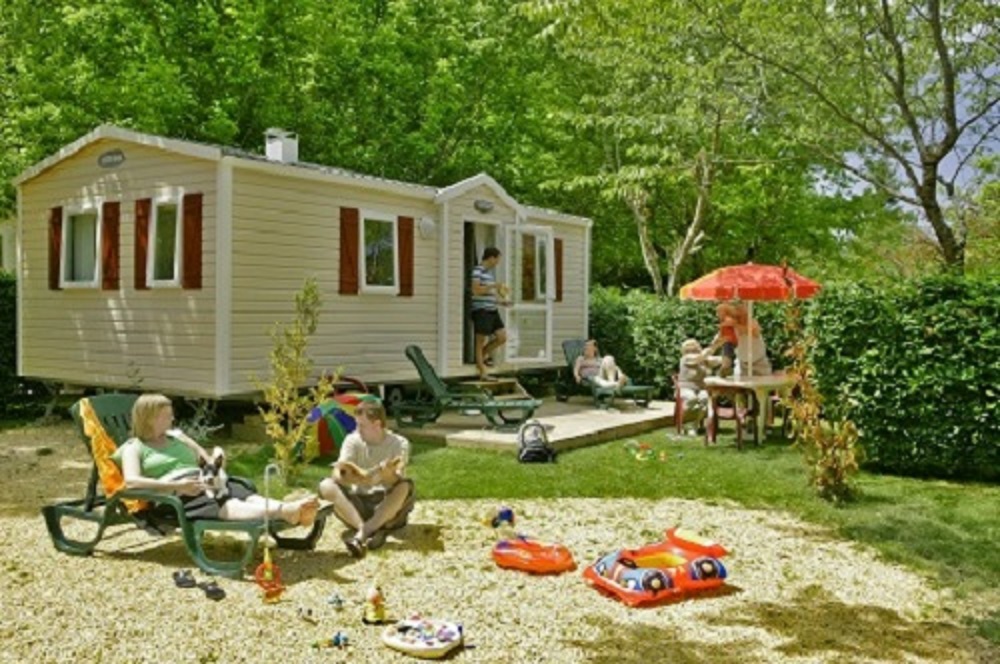 Wohnwagen mit Terrasse im Grünen, Gartenmöbel und Sonnenschirm, Spielzeug und Schwimmring auf dem Rasen.