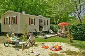 Wohnwagen mit Terrasse im Grünen, Gartenmöbel und Sonnenschirm, Spielzeug und Schwimmring auf dem Rasen.