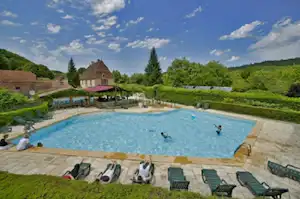 Großer Camping-Pool mit Liegen und Sonnenschirmen, umgeben von gepflegten Gärten und einem Steingebäude im Hintergrund.