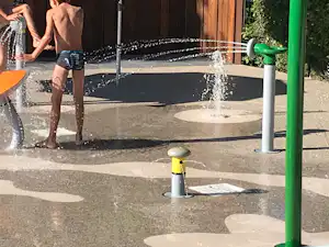 Spritzendes Wasser an einem Campingplatz-Brunnen, daneben ein Weg und ein gelb-schwarzer Poller vor einer Holzfassade.