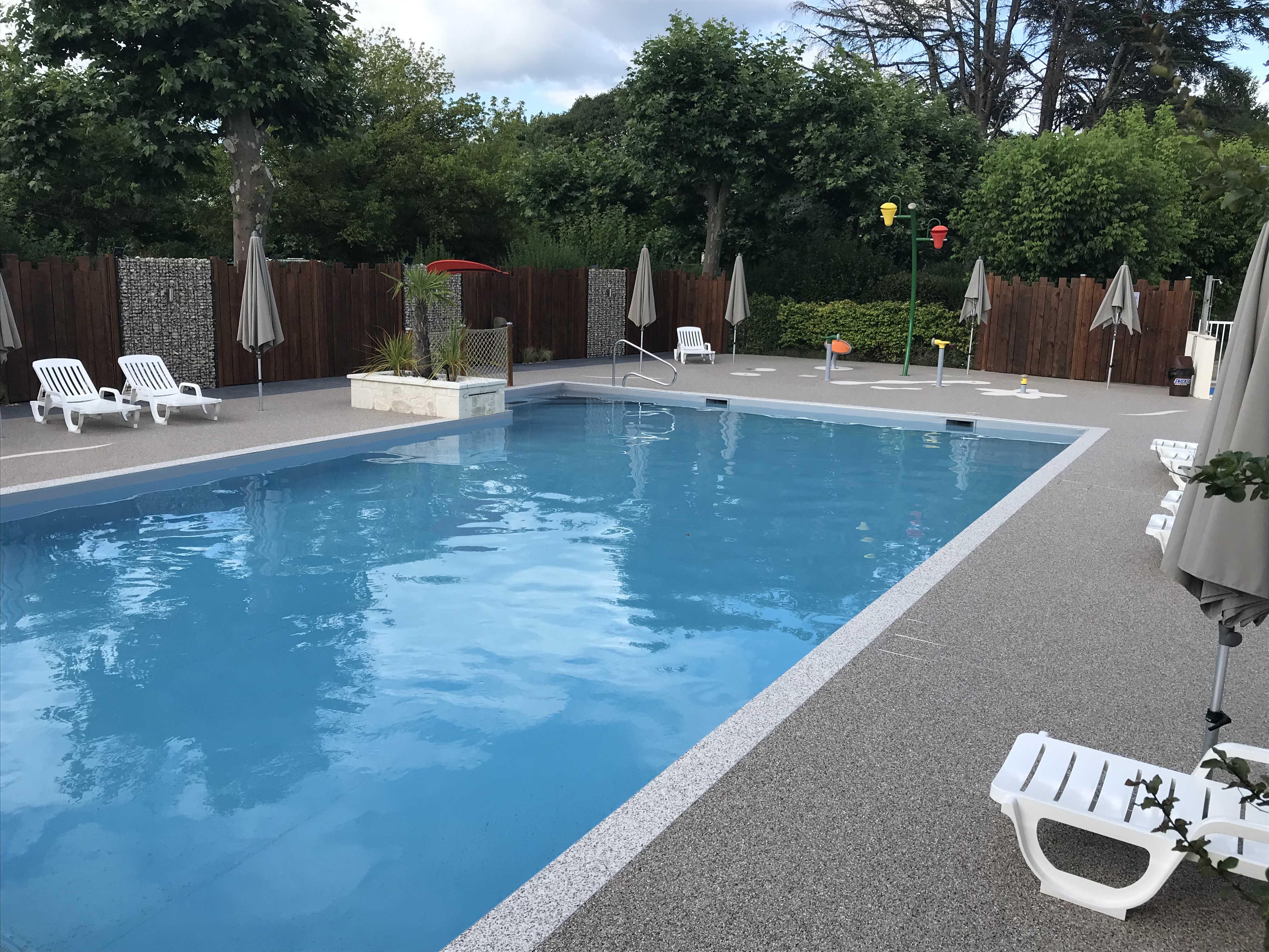 Blauer Camping-Pool mit Rand, weißen Liegen und Sonnenschirmen, umgeben von Hecken und Holzsichtschutz.