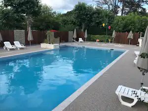 Blauer Camping-Pool mit Rand, weißen Liegen und Sonnenschirmen, umgeben von Hecken und Holzsichtschutz.