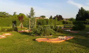 Grüne Campingwiese mit mehreren orangefarbenen Liegestühlen, Spielgeräten und Hecken im Hintergrund.