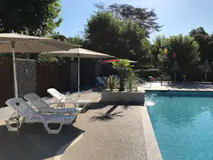 Pool am Campingplatz mit weißen Liegen, Sonnenschirm und Steinbeckenrand, umgeben von Bäumen und Sträuchern.