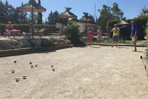 Familie auf dem Campingplatzweg vor Strohdächern, mit Spielgeräten und Liegen am Poolbereich, umgeben von Bäumen und Hecken.