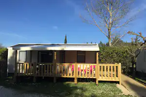 Bungalow mit Terrasse, Holzgeländer und Markise, im Garten mit Hecke und blühenden Blumen vor blauem Himmel.