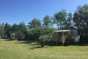 Mobilheim auf Stellplatz mit Holztreppe und Veranda, umgeben von Rasen und Bäumen im Campinggelände.