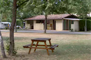Camping-Unterkunft mit Terrasse, Holzbank im Vordergrund und Bäumen im Garten, daneben ein Wohnwagen.