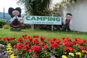 Camping-Schild „La Route Verte“ mit zwei Figuren, umgeben von Blumenbeeten und Rasen vor einer weißen Wand und Bäumen.