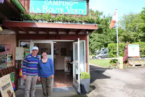 Empfangsbereich „ACCUEIL“ am Campingplatz mit zwei Personen vor dem Eingang, Schildern und Blumenbeeten im Grünen.
