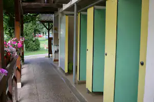 Bunte Spinde in einem überdachten Flur am Campingplatz, daneben Pflasterweg, Hecken und Blumenbeet im Hintergrund.