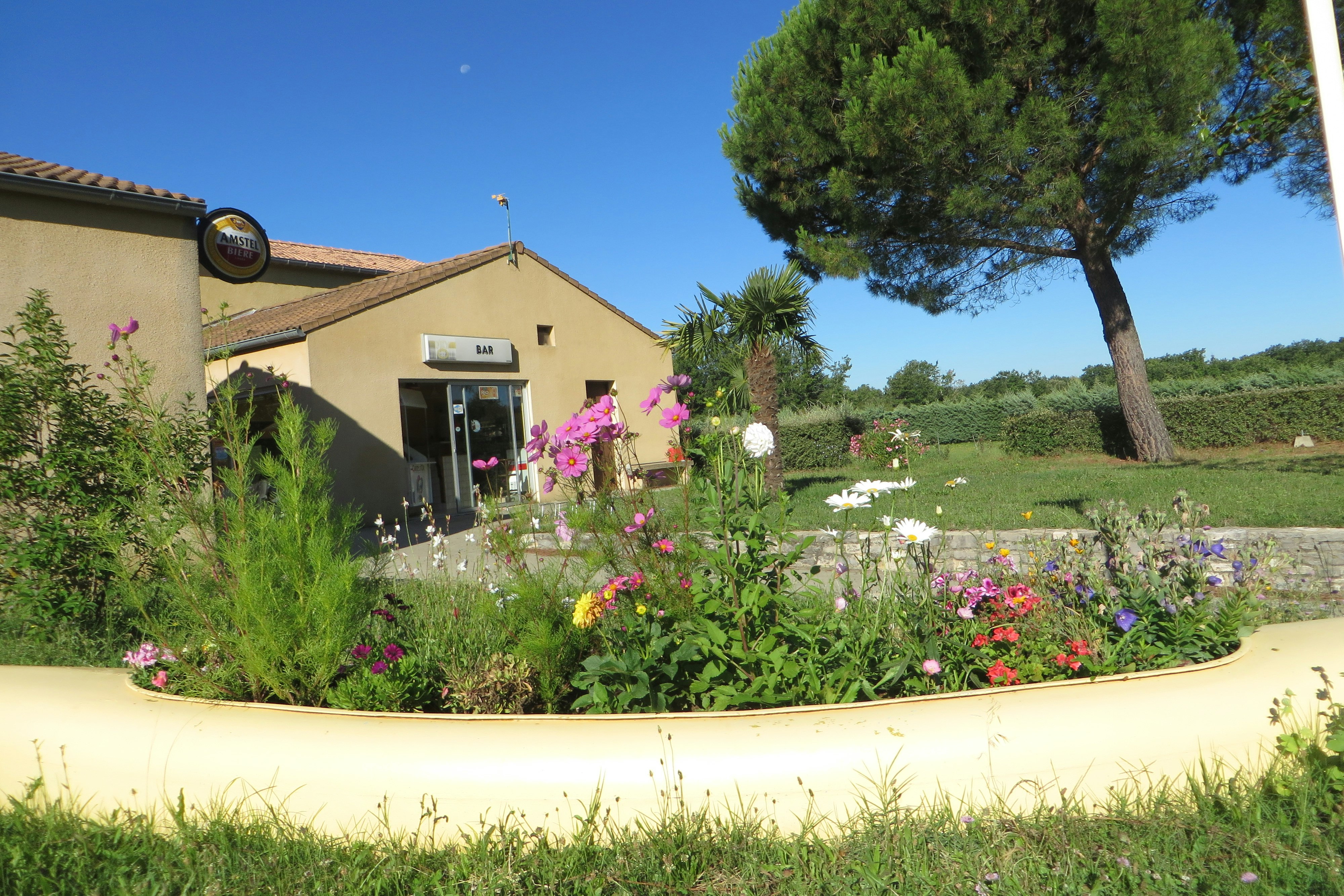 Campinghaus mit Eingangstür, davor ein Blumenbeet mit bunten Blüten und gepflegtem Rasen, im Hintergrund Bäume und Wiese.
