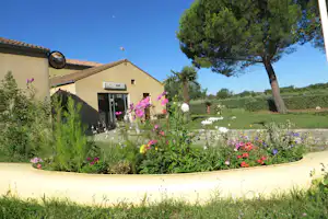 Campinghaus mit Eingangstür, davor ein Blumenbeet mit bunten Blüten und gepflegtem Rasen, im Hintergrund Bäume und Wiese.