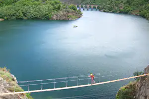 Hängebrücke über einem türkisfarbenen Fluss, im Hintergrund bewaldete Ufer und eine Steinbrücke mit Bögen.