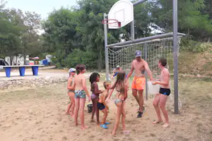 Kinder spielen am Basketballkorb auf dem Campingplatz, im Hintergrund Zelte, Bäume und ein Weg.