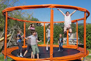 Kinder spielen in einer orangefarbenen Hüpfburg mit Netz und Polsterboden im Campinggarten, im Hintergrund Hecke und Bäume.