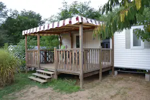Bungalow mit Holzterrasse und Streifen-Markise, seitlich weiße Fassade, Geländer und Bank vor dem Eingang.