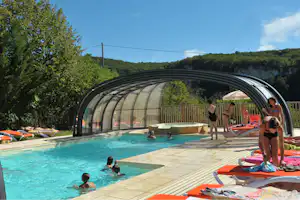 Überdachtes Camping-Poolbecken mit Liegen, Menschen im Wasser und am Rand, umgeben von Bäumen und Hügeln.