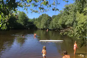Flussufer am Campingplatz: Kinder und Erwachsene schwimmen im ruhigen Wasser, daneben ein Paddelbrett auf der Oberfläche.