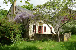 Ferienhaus mit roten Fensterläden und Gartenmauer, umgeben von grünem Rasen, Bäumen und blühenden Sträuchern.