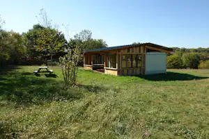 Holzveranda eines Mobilheims mit Vorhang, daneben ein Garten mit Rasen, Bäumen und einem Tisch.