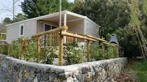 Mobilheim mit Veranda und Holzgeländer, umgeben von Hecken und einem niedrigen Steinwall am Campingplatz.