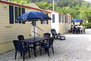 Bungalow mit Terrasse, Tisch und Stühlen unter zwei Sonnenschirmen, Kiesweg und Hecken im Hintergrund.