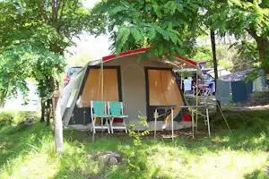 Campingzelt mit rotem Dach, davor Stühle und Tisch auf dem Gras, umgeben von Bäumen und Sichtschutz.