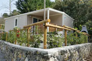 Bungalow mit Holzgeländer und Terrasse, umgeben von Sträuchern und Bäumen, dahinter ein Kiesweg und ein Steinsockel.