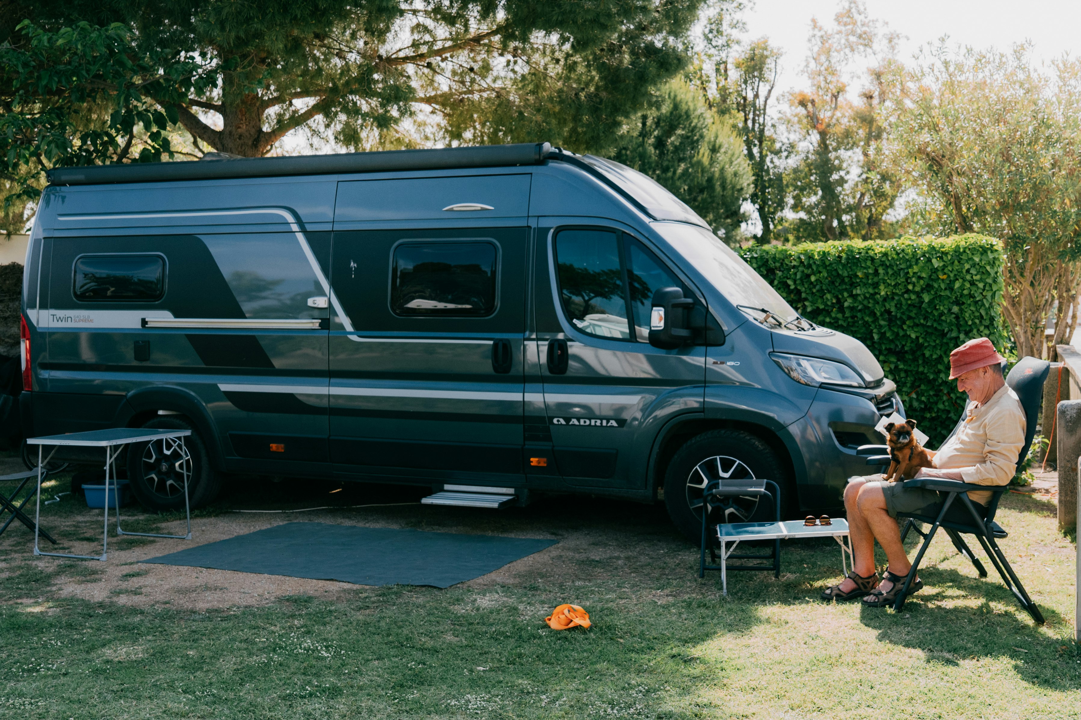 Grüner Camper-Van steht auf dem Stellplatz, daneben Tisch und Stühle; eine Person sitzt im Gartenbereich vor Hecke und Bäumen.