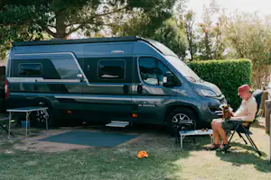 Grüner Camper-Van steht auf dem Stellplatz, daneben Tisch und Stühle; eine Person sitzt im Gartenbereich vor Hecke und Bäumen.