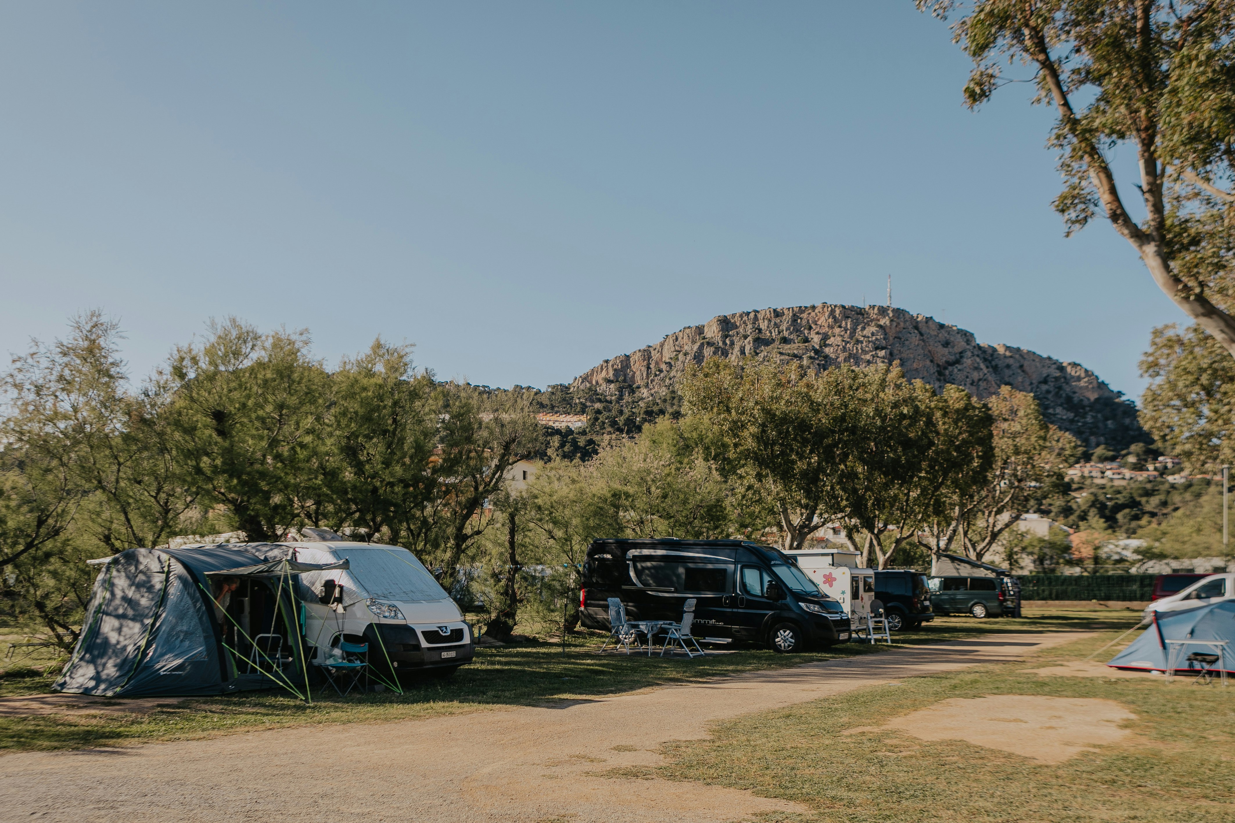 Campingplatz mit Zelten und Wohnmobilen, gepflasterter Weg und Bäume im Hintergrund vor einem bewaldeten Hügel.