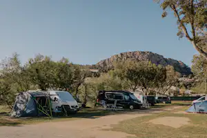 Campingplatz mit Zelten und Wohnmobilen, gepflasterter Weg und Bäume im Hintergrund vor einem bewaldeten Hügel.