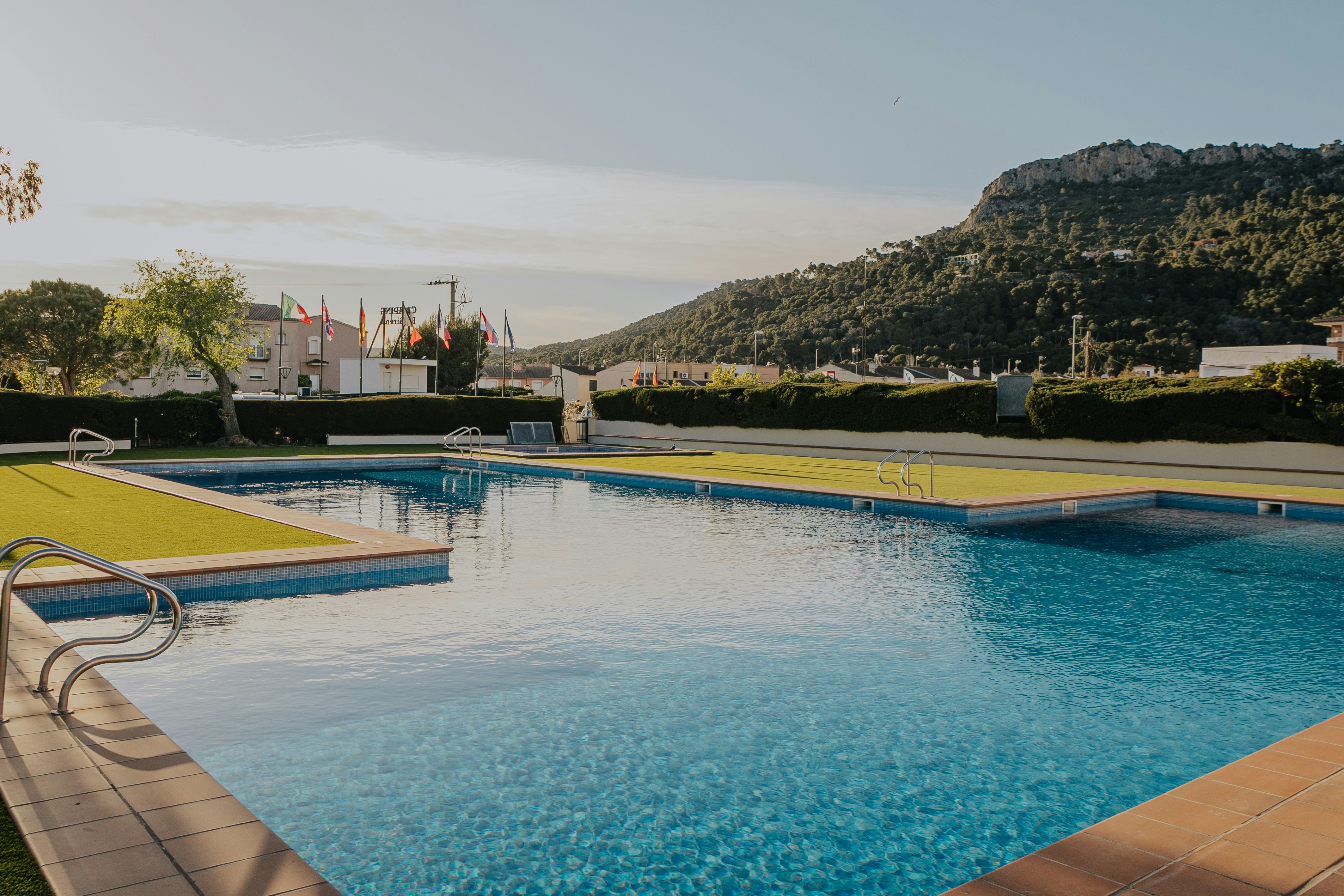 Schwimmbad mit blauem Wasser und Liegen am Rand, gepflasterte Terrasse und Hecken, im Hintergrund Gebäude und Hügel.
