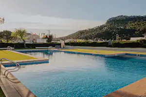 Schwimmbad mit blauem Wasser und Liegen am Rand, gepflasterte Terrasse und Hecken, im Hintergrund Gebäude und Hügel.