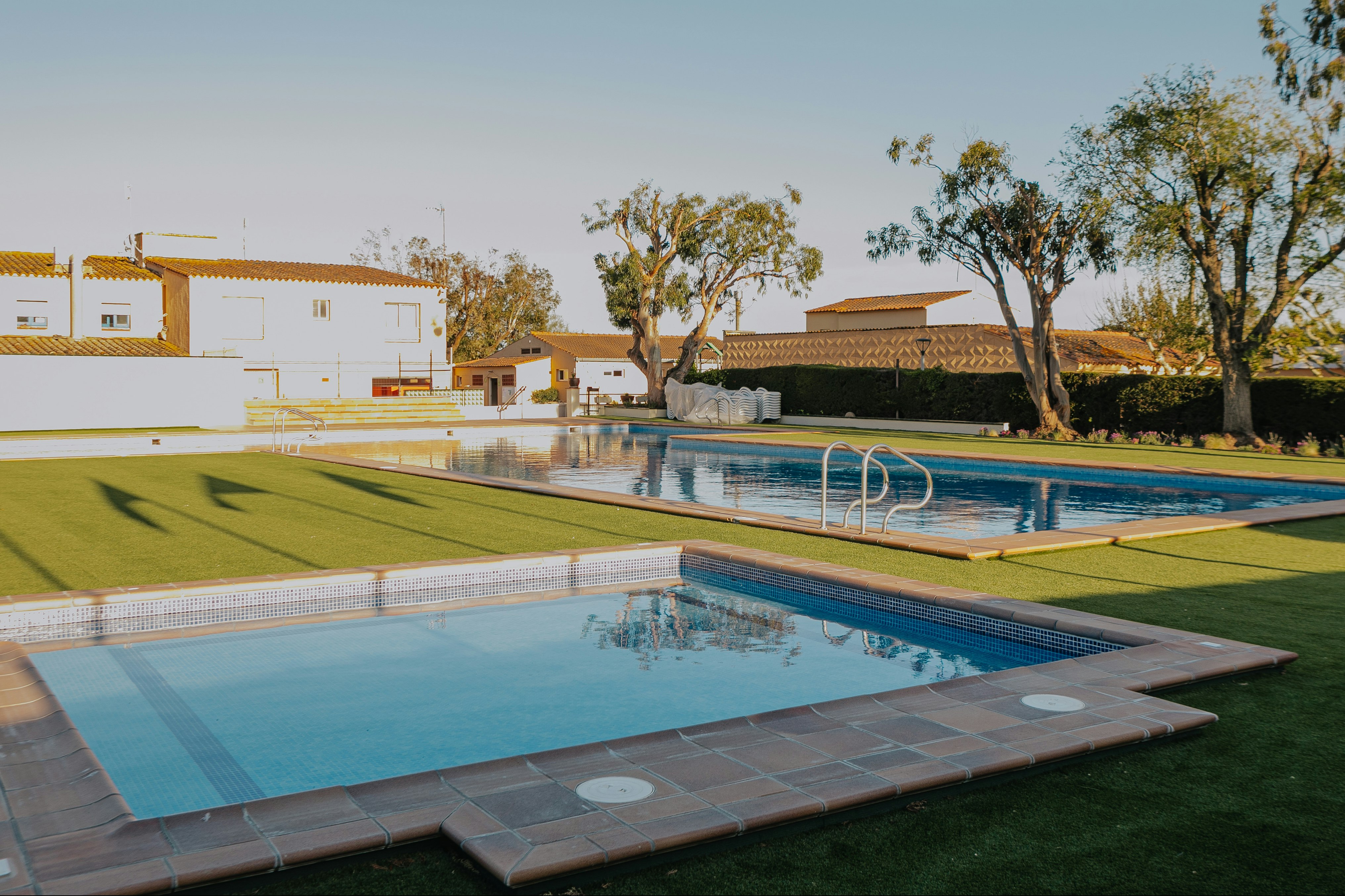 Schwimmbecken mit Sonnendeck und Rasenfläche, moderne Ferienanlage mit weißen Gebäuden und Bäumen im Hintergrund.