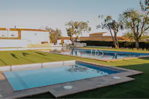 Schwimmbecken mit Sonnendeck und Rasenfläche, moderne Ferienanlage mit weißen Gebäuden und Bäumen im Hintergrund.