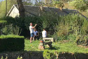Campinggarten mit Sitzbank aus Holz, Hecken und einem Haus im Hintergrund, im Vordergrund stehen zwei Personen.