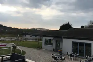 Campinghaus mit Terrasse, mehreren Stühlen und Tisch, Kiesweg und Rasenfläche im Garten, im Hintergrund weitere Gebäude.