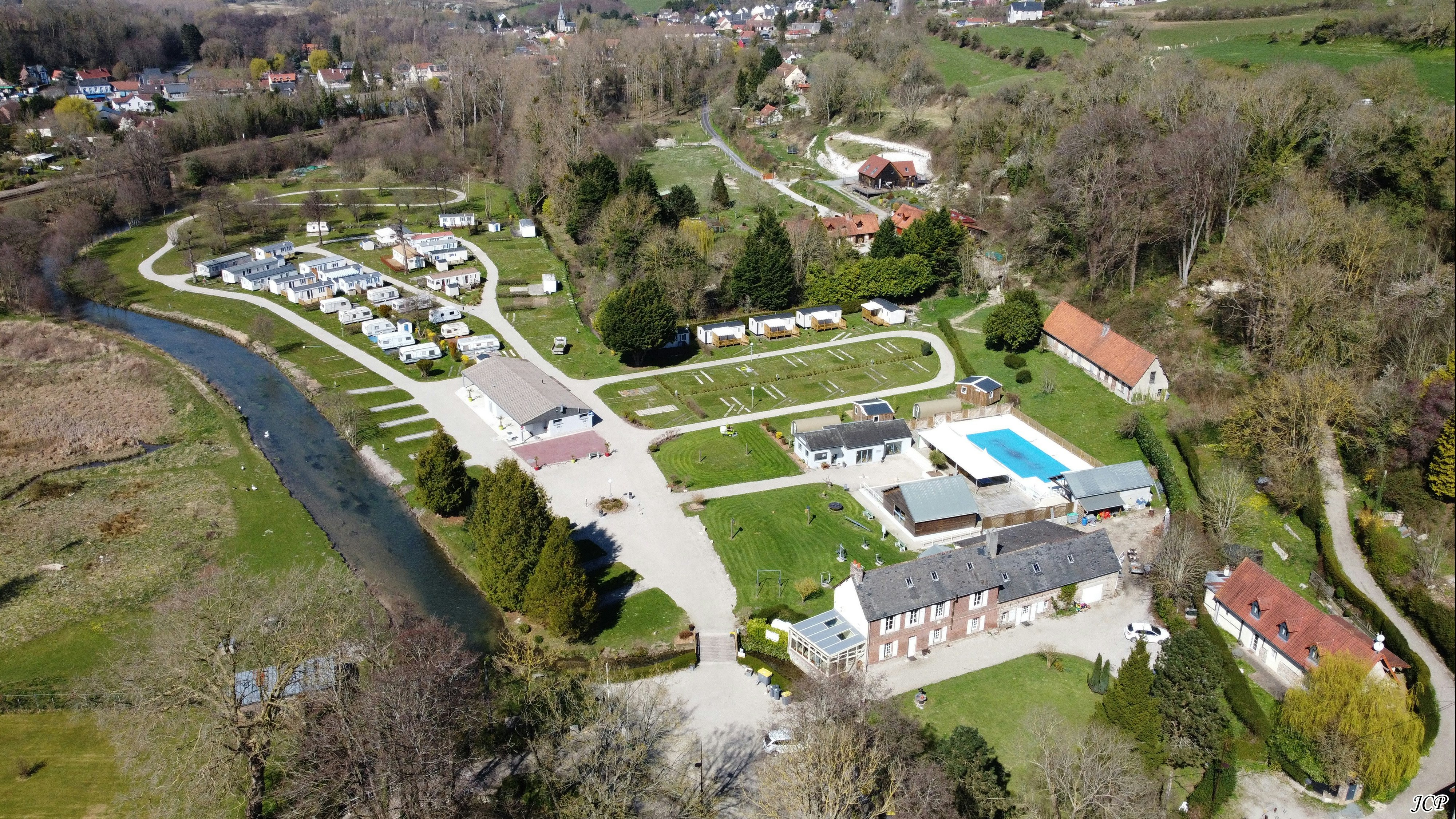 Luftaufnahme eines Campingplatzes mit Stellplätzen, Wohnmobilen, Pool und Wegen zwischen Bäumen und Wiesen.