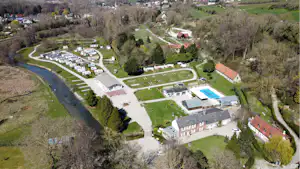 Luftaufnahme eines Campingplatzes mit Stellplätzen, Wohnmobilen, Pool und Wegen zwischen Bäumen und Wiesen.
