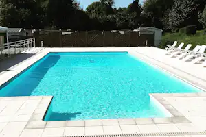 Schwimmbecken auf dem Campingplatz mit weißen Liegen, gepflasterter Terrasse und Zaun im Hintergrund, umgeben von Bäumen.