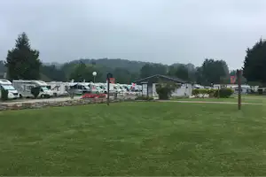 Grüner Rasen auf einem Campingplatz mit Stellplätzen für Wohnmobile und Wohnwagen, Bäume und Wege im Hintergrund.