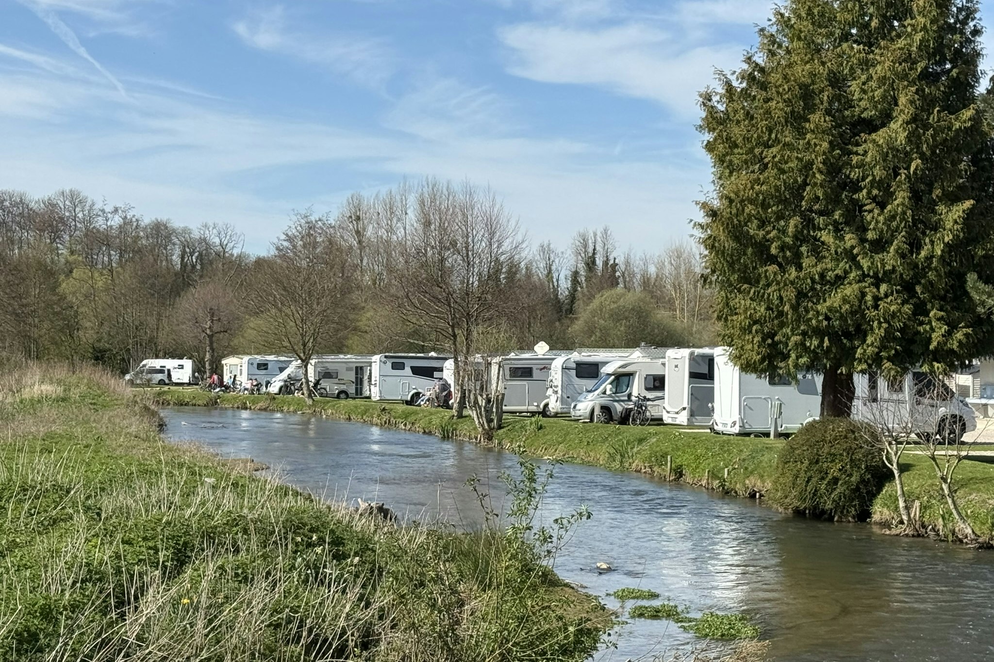Reihe von Wohnwagen am Campingplatz entlang eines kleinen Flusses, mit Gras- und Ufervegetation sowie Bäumen im Hintergrund.