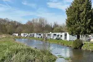 Reihe von Wohnwagen am Campingplatz entlang eines kleinen Flusses, mit Gras- und Ufervegetation sowie Bäumen im Hintergrund.